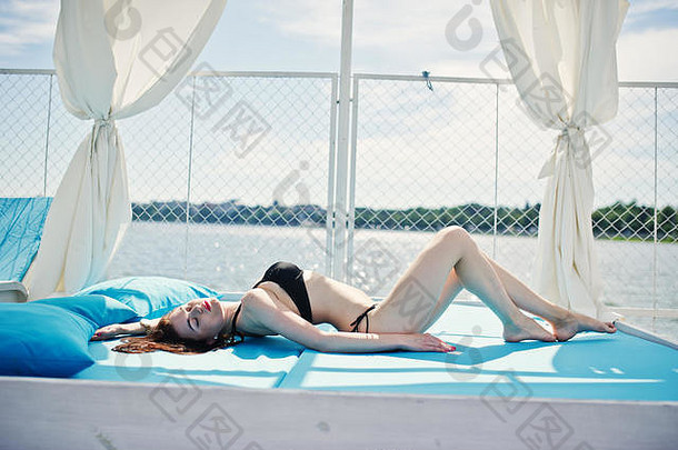 一个穿着黑色<strong>比基尼泳衣</strong>的美丽女孩躺在湖边的躺椅上的肖像。