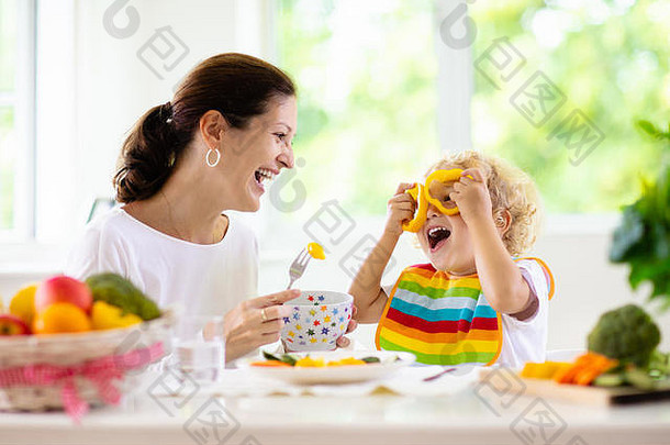 母亲给孩子喂蔬菜。妈妈在有窗户的白色厨房里喂孩子。坐在高脚椅上的，吃着健康的蒸胡萝卜和土豆午餐