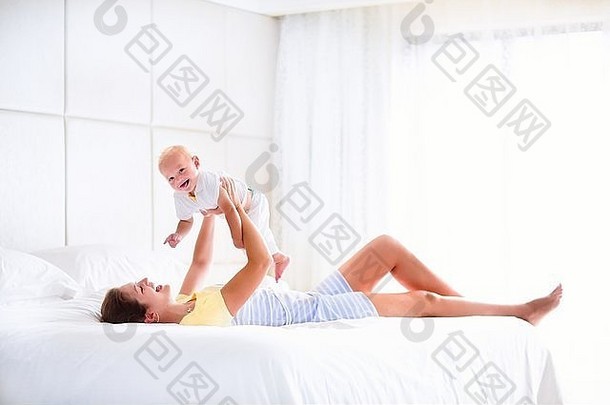年轻快乐的母亲抱着她可爱的孩子，可爱的金发卷发男孩，在白色的床上玩耍，享受着一个阳光明媚的早晨