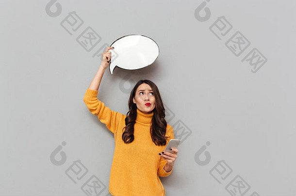 体贴的黑发女人，穿着毛衣，头顶上挂着空白的语音泡泡，手持智能手机，在灰色背景下抬头看