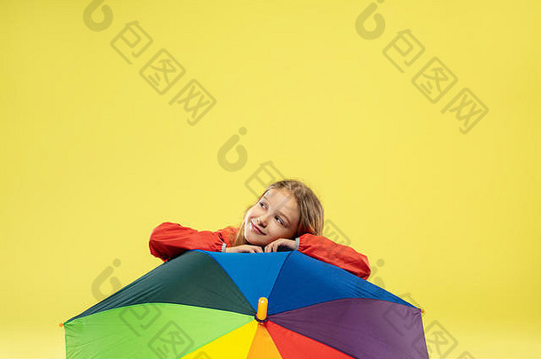 黄色摄影棚背景上，一位穿着雨衣、带彩虹色雨伞的时尚女孩的全长肖像。看起来很高兴。儿童秋季和春季时装。可爱时尚的金发女孩。