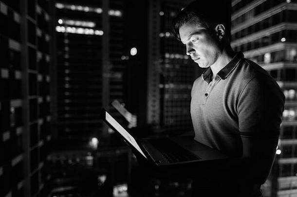 年轻英俊的男子用笔记本电脑对着玻璃窗，夜晚可以看到城市的景色