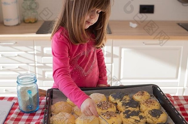 高加索女孩，3岁，在烹调前在烤箱里的面包包子上撒种子。封锁活动的想法。