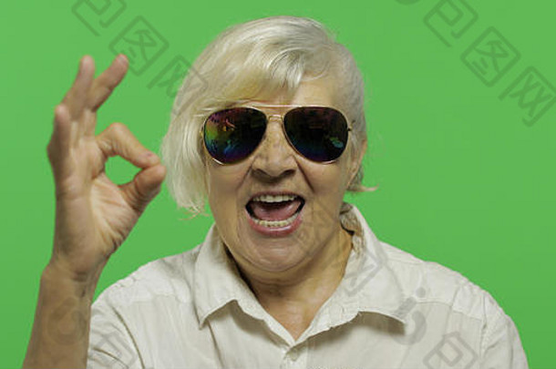 一位戴太阳镜的老太太摆出一副很好的姿势，微笑着。穿着白衬衫的漂亮老奶奶。放置您的徽标或文字。色度键。绿色屏幕背景