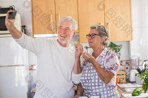 快乐美丽的老年夫妇一起在厨房做午餐。这名男子将自拍带到双方，以获得乐趣