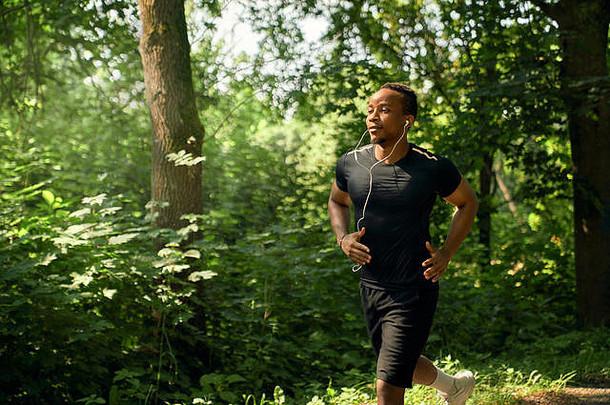 夏天的早晨，一个非洲男人在花园里跑步，他有着<strong>运动</strong>的身体。穿着黑色t恤和短裤的<strong>运动</strong>员戴着耳机听音乐。跑步训练。
