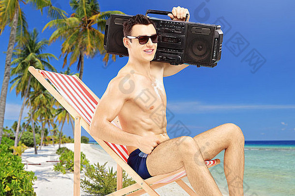 一个帅哥在一张椅子上放松，肩上扛着收音机，在一个热带海滩上，海边和棕榈树旁
