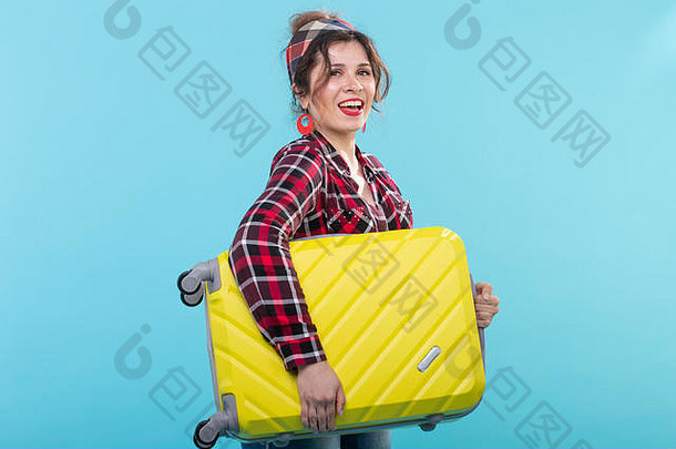 身着格子衬衫、手持黄色手提箱、在蓝色背景上摆姿势的积极微笑的年轻女子。旅游与旅游的概念