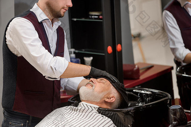 无法辨认的理发师正在用毛巾在黑色洗脸盆里擦拭客人的头部。男理发师为<strong>老客户</strong>的头发定型，留着灰胡子。在理发店工作的发型师。