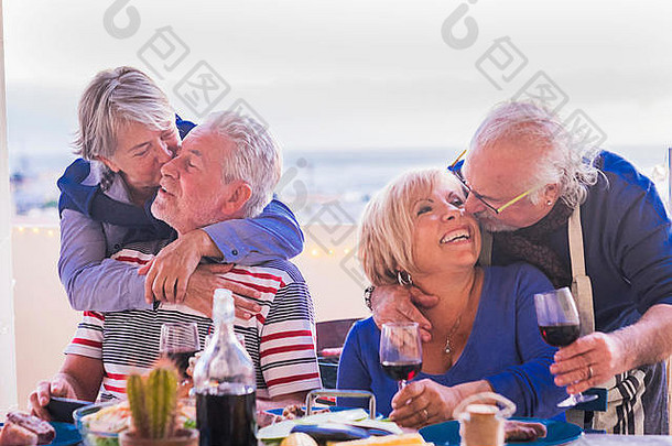 4位白发老人坐在阳台上的桌子旁拥抱亲吻。背景是黄色的灯光和海洋。面带微笑