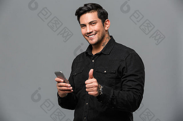 照片微笑年轻的男人。站孤立的灰色墙相机显示拇指聊天移动电话