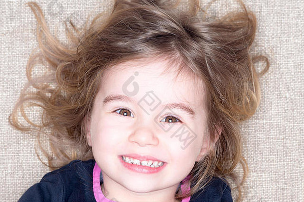 三岁小女孩躺在地毯上，头发蓬乱，脸上带着幸福的表情对着镜头咧嘴笑