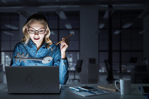 迷人的金发女郎戴着眼镜在黑暗的办公室里使用笔记本电脑。混合介质