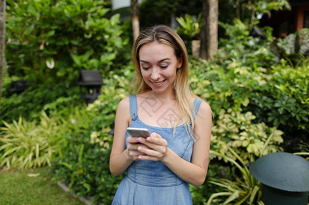 年轻的美国女人聊天智能手机花园树穿牛仔裤背心裙