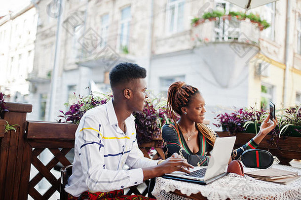 一对英俊时尚的非洲裔美国夫妇坐在户外咖啡馆里，拿着笔记本电脑，用手机自拍。