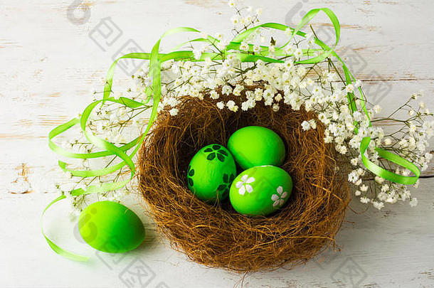 绿色复活节彩蛋，绿色缎带，白色婴儿呼吸花，白色木质背景，鸟巢，俯视图