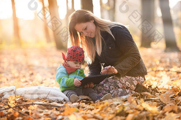 母亲和坐在公园里的毯子上，使用数字平板电脑。年轻的妈妈教他的孩子如何使用带触摸屏的平板电脑。小孩。