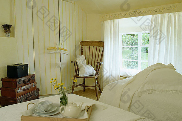 奶油乡村卧室床上的茶壶和茶杯托盘，窗户上有白色窗帘，衣柜门上有黄色条纹