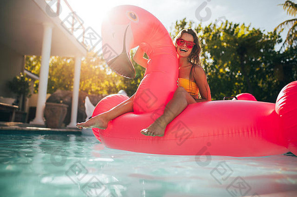 年轻女子在一个充气的火烈鸟游泳池浮动床垫上嬉笑。夏日游泳池里迷人的女人。