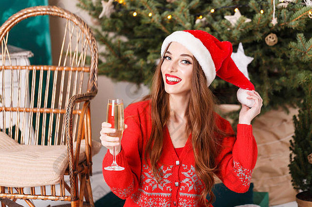 穿着圣诞帽和红色毛衣的迷人圣诞女郎，手拿帽子坐在舒适的新年云杉旁的地板上，手拿香槟、新年、圣诞节、假日、纪念品、礼品、购物、折扣、商店、雪人圣诞老人、化妆、发型、狂欢节。