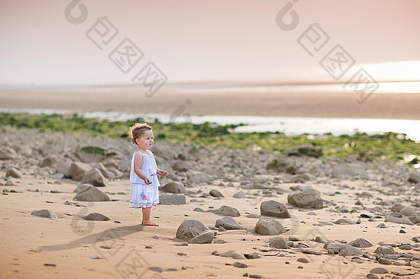 美丽的<strong>小</strong>，<strong>卷发</strong>，穿着白色连衣裙，日落时在海滩上散步