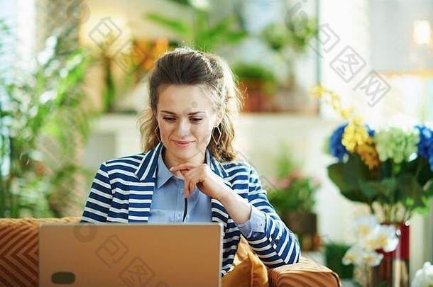 沉思的时尚的女人蓝色的上衣条纹夹克坐着沙发上移动PC现代房子阳光明媚的一天