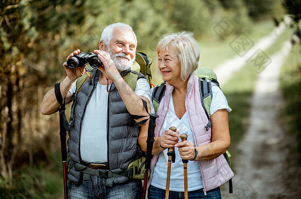 一对幸福的老年夫妇带着双筒望远镜、背包和<strong>登山杖</strong>站在一起在森林里徒步旅行的照片。退休后积极生活方式的概念