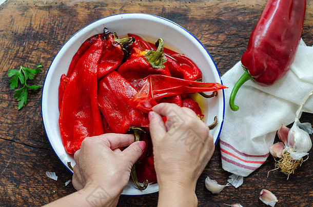 木桌上的一个碗里放着有机腌制的烤红辣椒。女人一只手拿着烤红辣椒，另一只手在剥胡椒皮