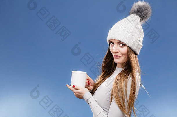 一名女子戴着冬帽，身穿灰色运动保暖内衣，用于滑雪训练。她拿着一个盛着茶或咖啡的杯子，在蓝色的摄影棚里为自己取暖。