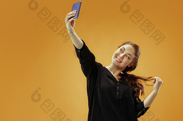 一个自信、快乐、微笑的休闲女孩在金色背景下用手机拍摄自拍照片。
