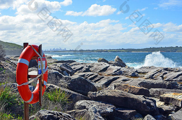 澳大利亚新南威尔士州，岩石海岸线上的岩石平台上有救生圈，背景是破碎的海浪和悉尼市的天际线。
