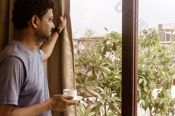 一个男人端着一杯香喷喷的咖啡，放松地看着窗外。早晨的阳光从外面射进来。周末快乐