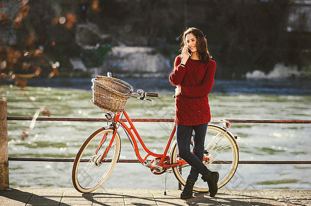 在巴塞尔市的一条河边，一位年轻的女游客站在一辆<strong>自行车</strong>旁边，正在享受这一乐趣。在瑞士旅行。年轻的白人妇女在说话
