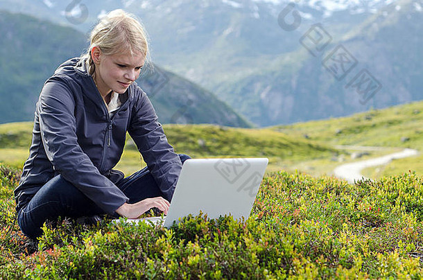 在高山景观前的高山草甸上，一位年轻的金发女郎拿着笔记本电脑坐着