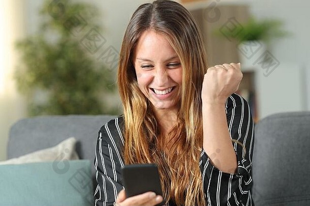 兴奋的女人坐在家客厅的沙发上查看手机在线内容庆祝好消息