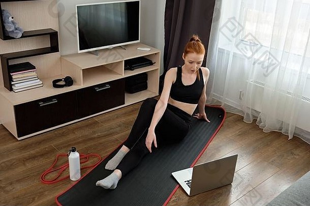 一个穿着运动服的女孩坐在笔记本电脑前的垫子上，坐在上面的框架上，在家里健身