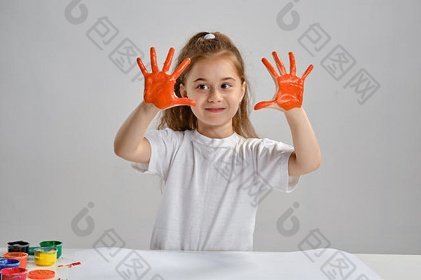 穿着白色t恤的小女孩坐在桌旁，手里拿着<strong>惠</strong>特曼和五颜六色的颜料，展示着她画过的手。白色的隔离带。中等特写镜头。