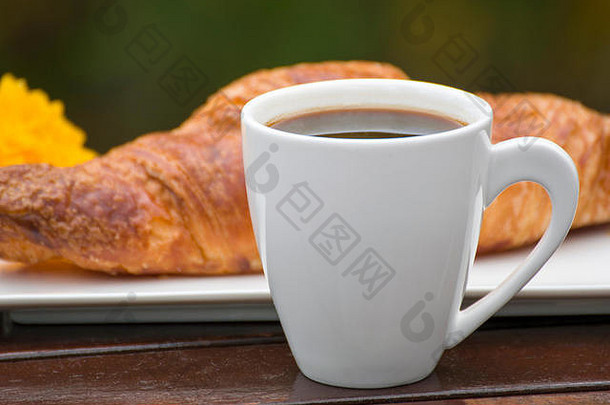 一杯黑咖啡和一个牛角面包放在木桌上的特写镜头，外面。