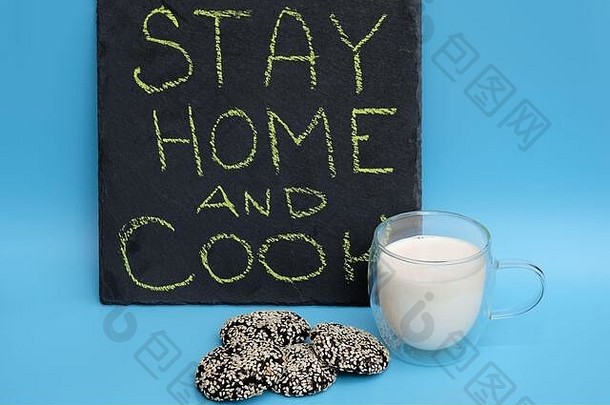 蓝色背景上的一杯牛奶和巧克力饼干。黑黑板上的铭文留在家里做饭。概念-检疫，wh