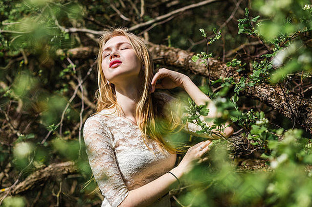 春天，在背景花树上，一位留着长发、身穿白色连衣裙的美丽金发女郎的肖像，闭着<strong>眼睛</strong>