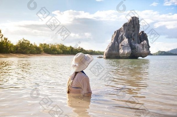 一个戴着白帽子的年轻女孩正坐在热带岛屿上的水中享受假期。暑期和旅游度假概念