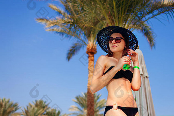 一位身穿比基尼、头戴帽子的女士拿着鸡尾酒对着棕榈树。<strong>暑假</strong>一<strong>价</strong>全包