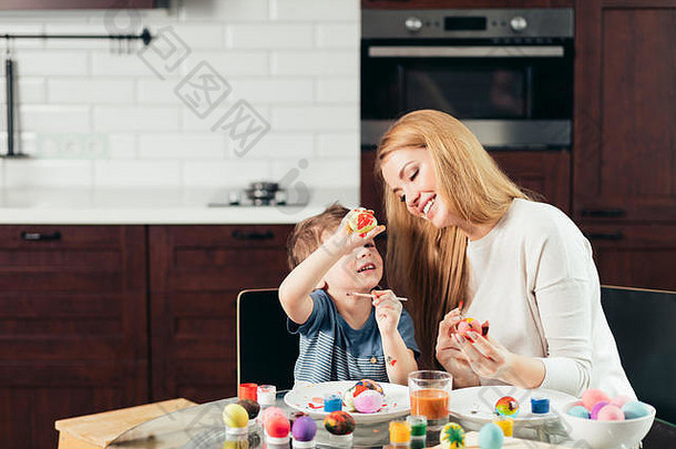 年轻漂亮的金发妈妈和她可爱的四岁儿子在家里的厨房里画复活<strong>节</strong>彩蛋时玩得很开心，面带微笑，培养了创造力