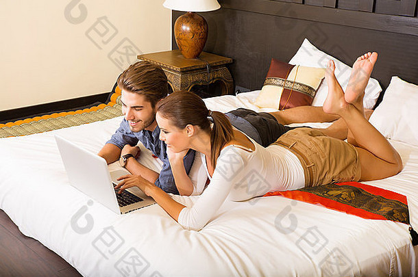 一对年轻夫妇躺在床上，在亚洲酒店房间里用笔记本。