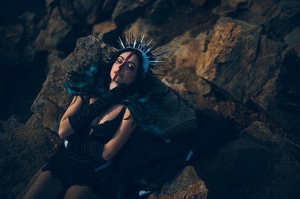 一个年轻女子，形象是一个仙女和一个女巫，穿着黑色衣服，头戴皇冠躺在岩石上。