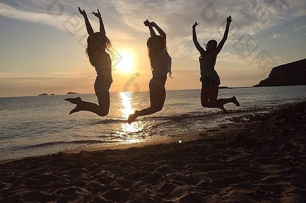 日落时三个年轻女孩在海滩上跳跃的剪影