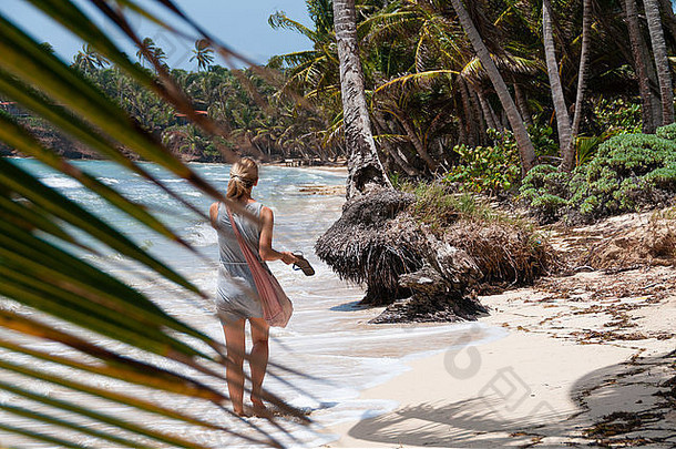 美丽的金发女人，带着粉红色的袋子，光着脚在小小的<strong>玉米</strong>岛上孤独的加勒比海<strong>白</strong>色沙滩和椰子树后面散步