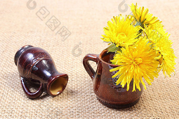 一束黄色的蒲公英鲜花在棕色花瓶里，背景是一个翻倒的花瓶，躺在黄麻帆布上