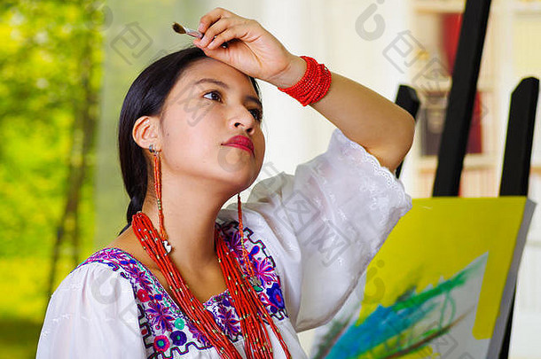 身穿安第斯传统服装的年轻黑发女子，手持画笔，用手擦拭额头，在工作室内，以花园窗户为背景