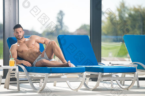 年轻人在游泳池旁放着橙汁的日光浴椅上休息